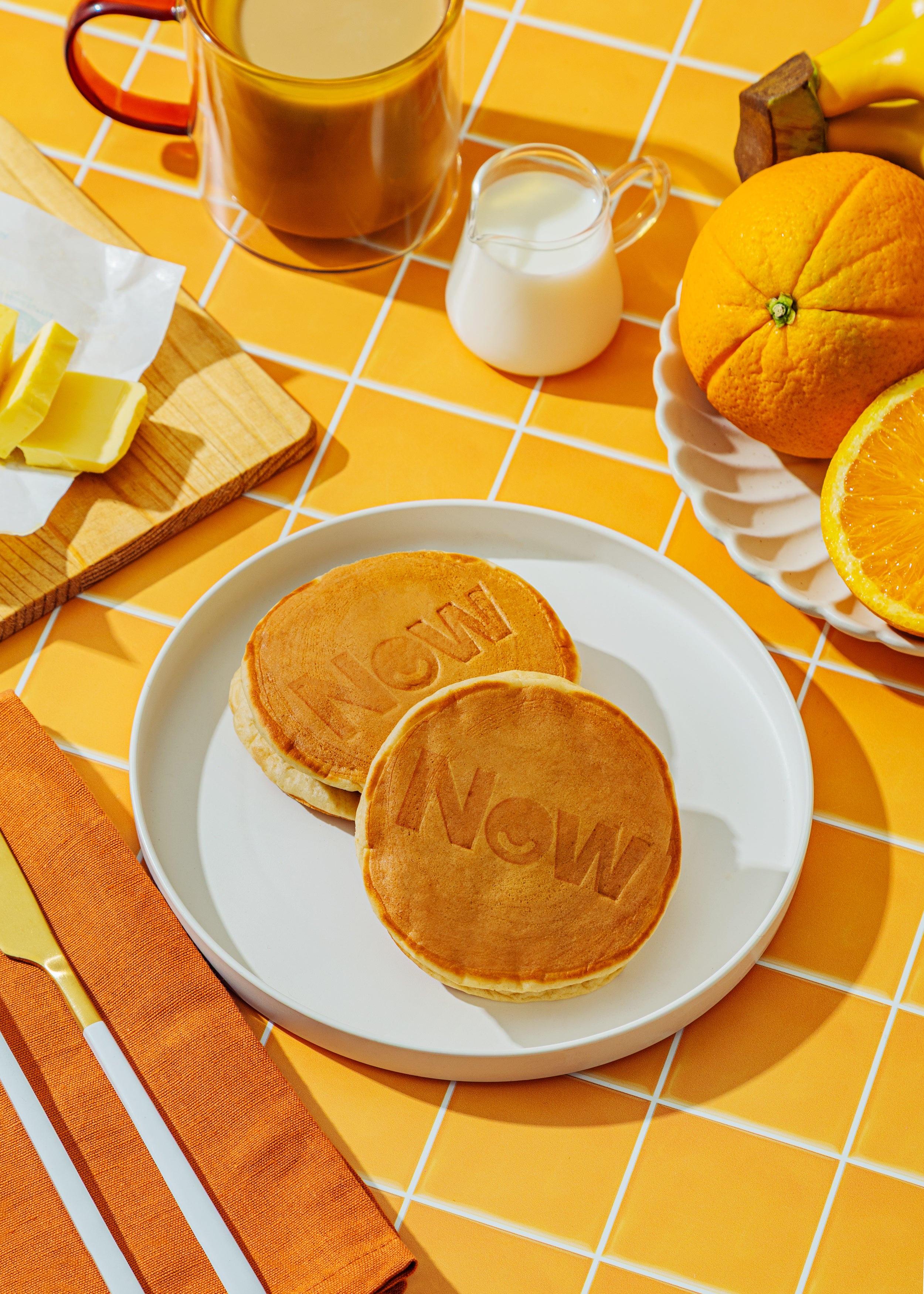 Pancakes with 'Now' text on a plate, served with orange juice and milk on a yellow checkered tablecloth.